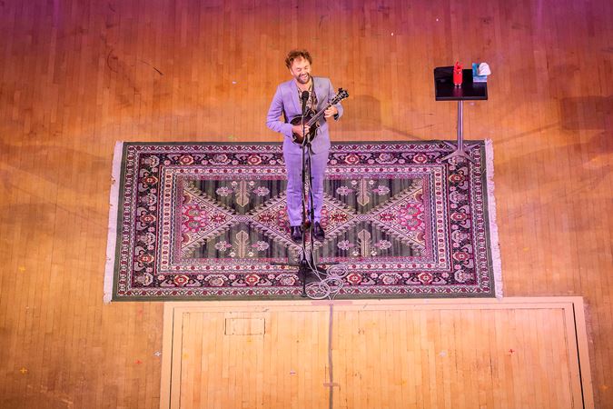 Chris Thile sings and plays mandolin, standing in the middle of an ornate, patterned carpet rolled out for him on stage